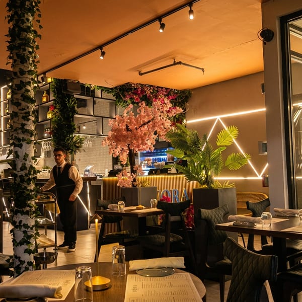 A restaurant interior with a waiter, tables set for dining, and decorative plants and lighting.