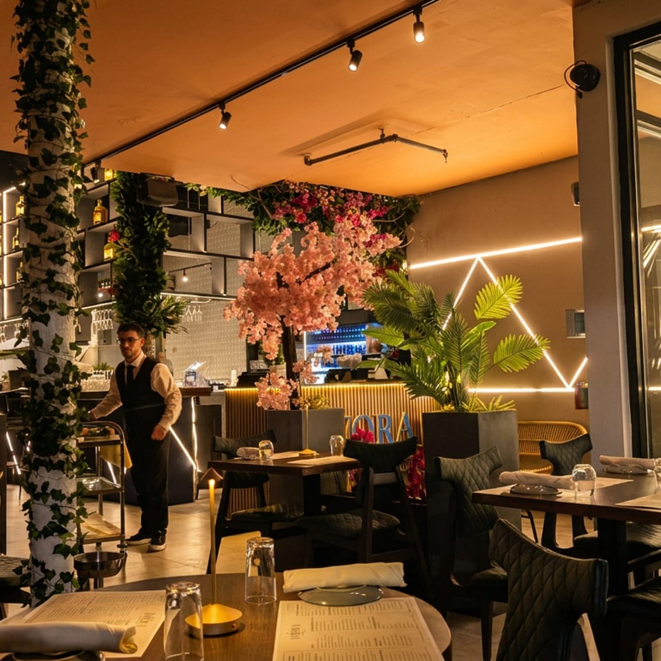 A restaurant interior with a waiter, tables set for dining, and decorative plants and lighting.