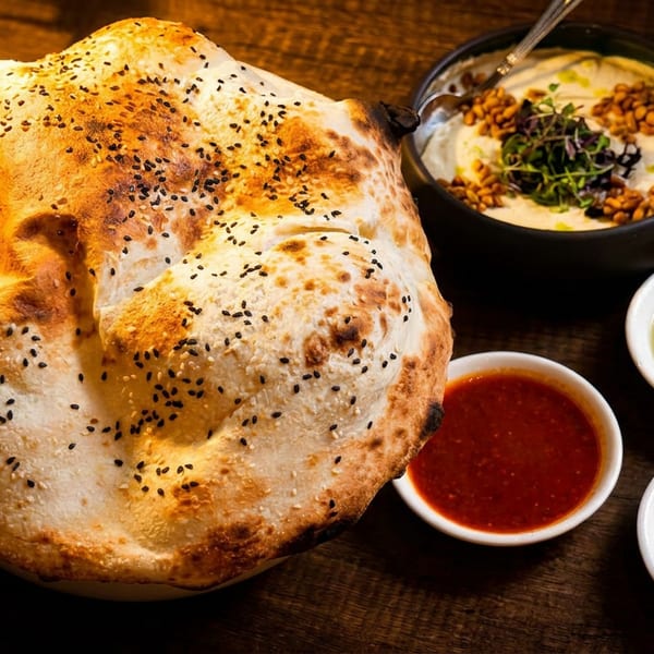 Freshly baked flatbread with sesame seeds, hummus, olive oil, red sauce, and white sauce on a wooden table.