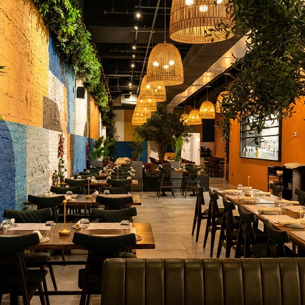 A long, modern restaurant dining room with colorful walls, wicker pendant lights, and lush greenery.